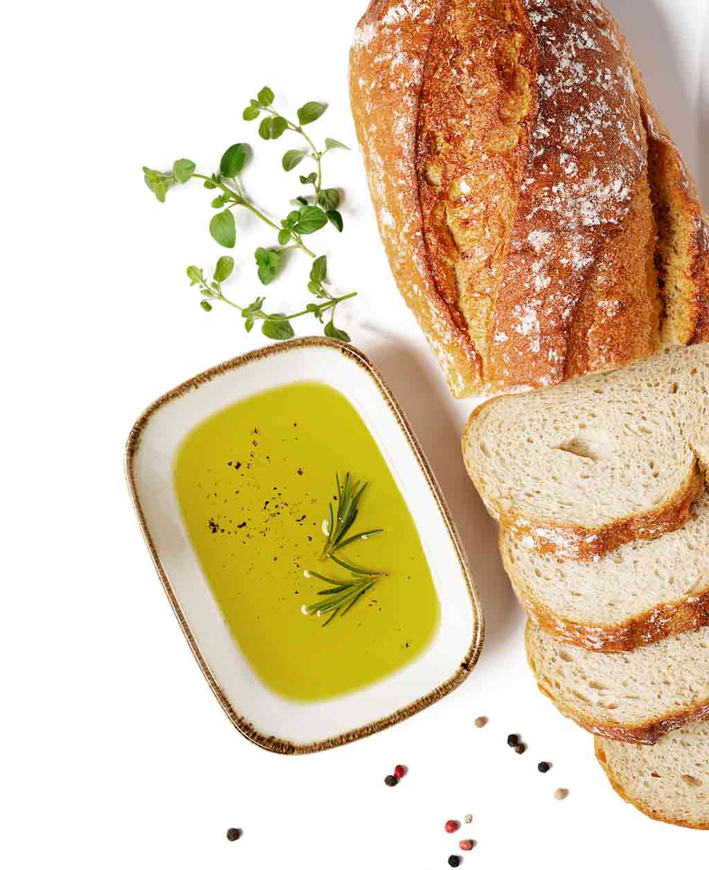 In der Mitte steht ein rechteckiges Schälchen gefüllt mit Olivenöl und ein bisschen Pfeffer und Rosmarinzweigen. Daneben liegt ein Baguette zum Teil in Scheiben geschnitten. Ein paar Kräuterzweige liegen über dem Schälchen. Das Foto hat einen weißen Hintergrund